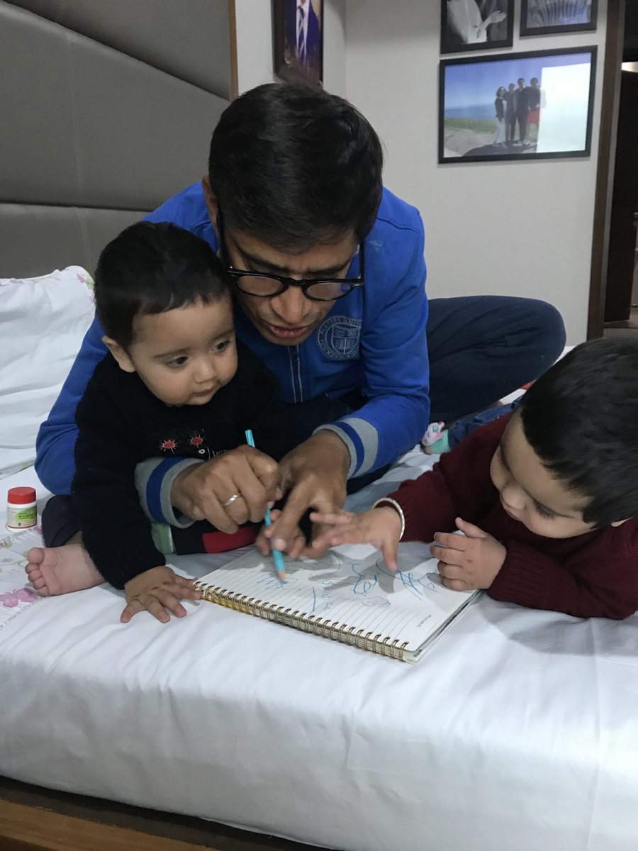 एक पिता notebook पर लिखते हुए, दो छोटे बच्चे उनके pencil को ध्यान से देख रहे हैं