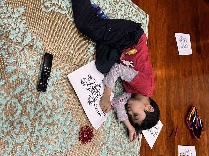 ऊपर से लिया गया photo — एक बच्चा ज़मीन पर लेटकर colouring कर रहा है, आस-पास कई पूरी की हुई sheets बिखरी हैं